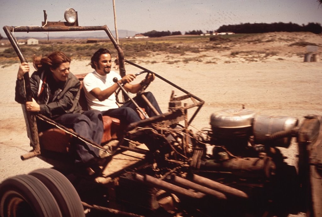 A grinning young man drives a homemade open-frame dune buggy across the sand in 1972 while a young woman rides beside him, her hair blowing