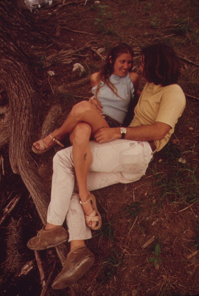 A young couple sitting close together on tree roots beside the Frio River in 1973, she in a cherry-print halter top, smiling at each other