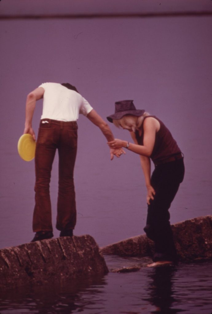 A young man holding a yellow Frisbee steadies a barefoot young woman by the hand as she steps across shoreline rocks in Lincoln Park, Chicago, in 1973