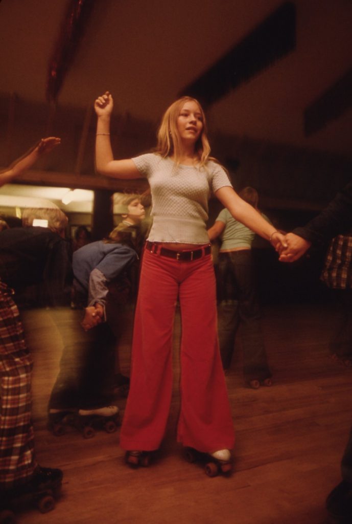 A teenage girl in red corduroy bell-bottoms roller skates while holding a hand that reaches in from the edge of the frame, at a Minnesota roller rink in 1974