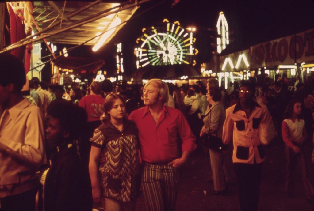 A young couple walking the state fair midway at night in 1972, a lit Ferris wheel and game booths glowing behind them