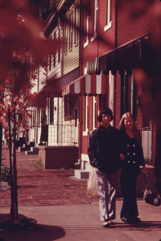 A young couple walking arm in arm down a Chicago sidewalk past striped awnings and autumn leaves in October 1974