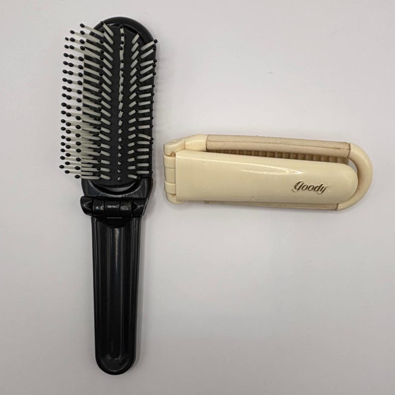 An open black Goody folding hairbrush beside a closed cream-colored Goody brush on a white background