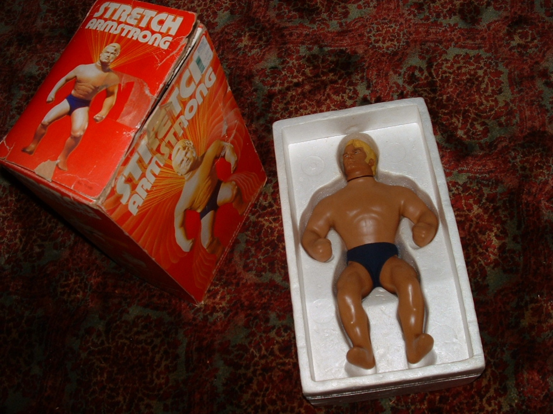 A vintage Stretch Armstrong figure in its foam tray beside the red original box
