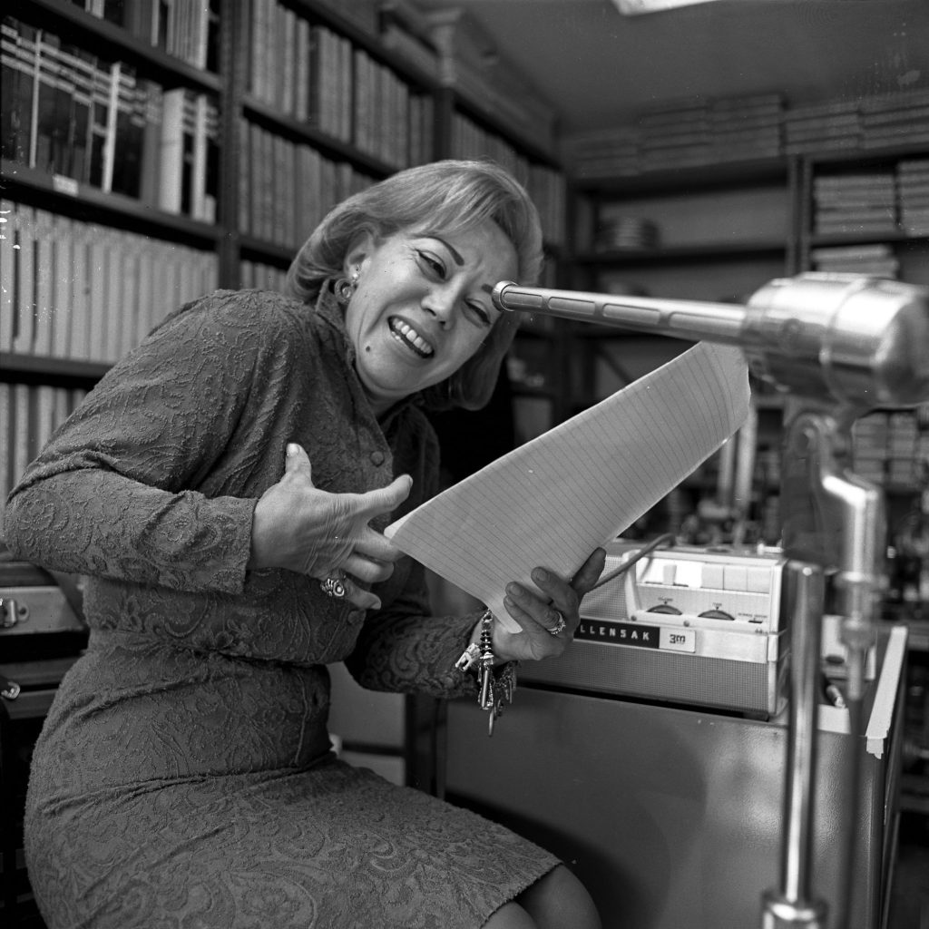 June Foray, seated in a patterned dress, grimaces comically while reading from a script into a studio microphone in front of shelves of books and a tape recorder.