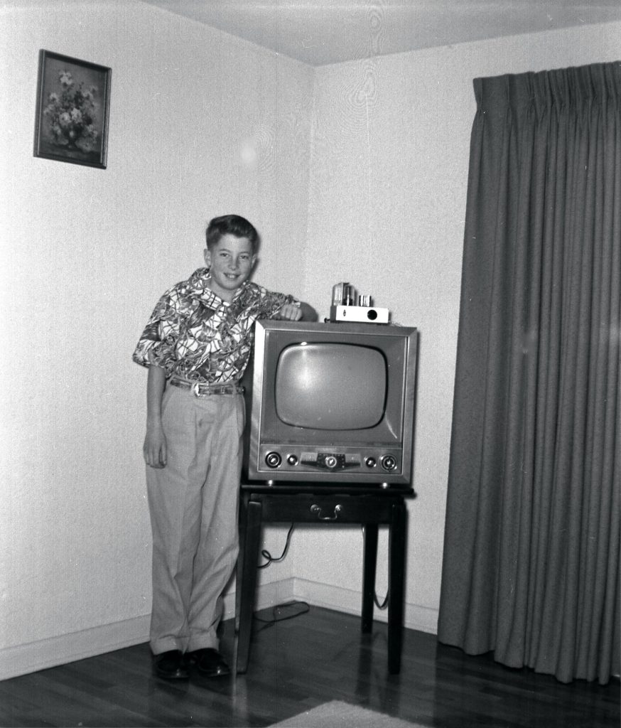 A smiling boy in a patterned short-sleeved shirt stands with his arm resting on a small table-model television set in the corner of a living room.