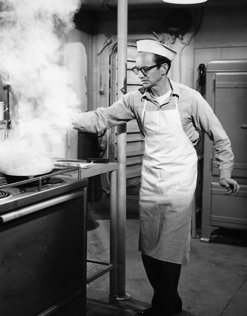 Wally Cox in glasses, a white sailor's cap and an apron reaches toward a stove where a frying pan sends up a large cloud of steam.