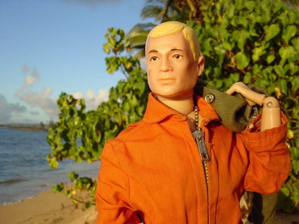 A 12-inch G.I. Joe figure with painted blond hair in an orange flight suit, a green duffel bag over his shoulder, posed on a beach