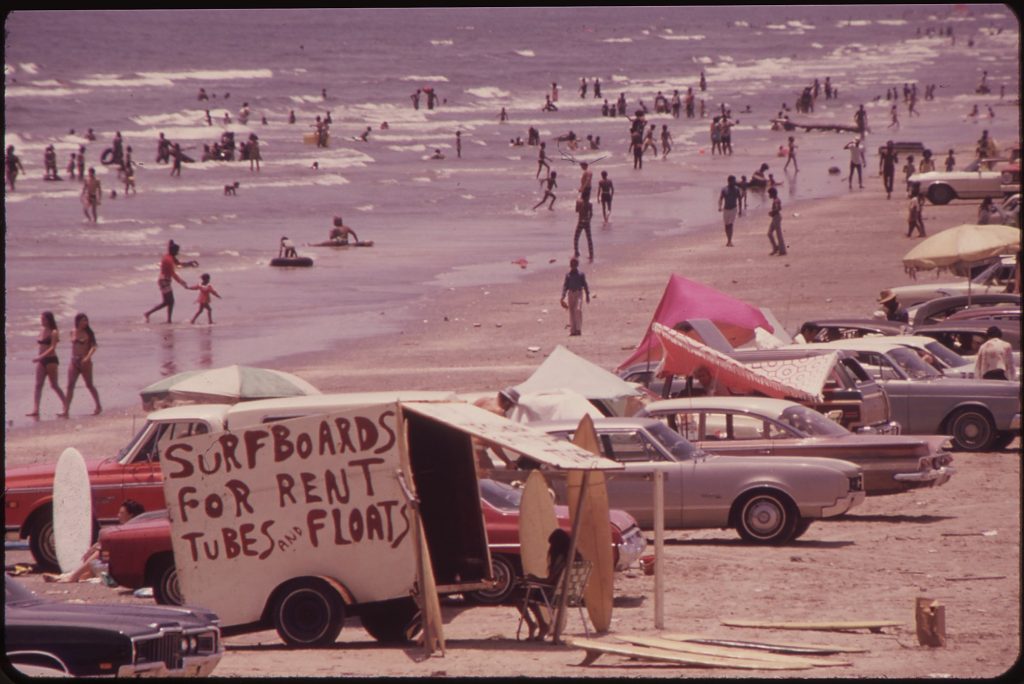 A crowded beach with swimmers in the surf, cars parked on the sand, and a homemade sign on a trailer reading Surfboards for Rent, Tubes and Floats.