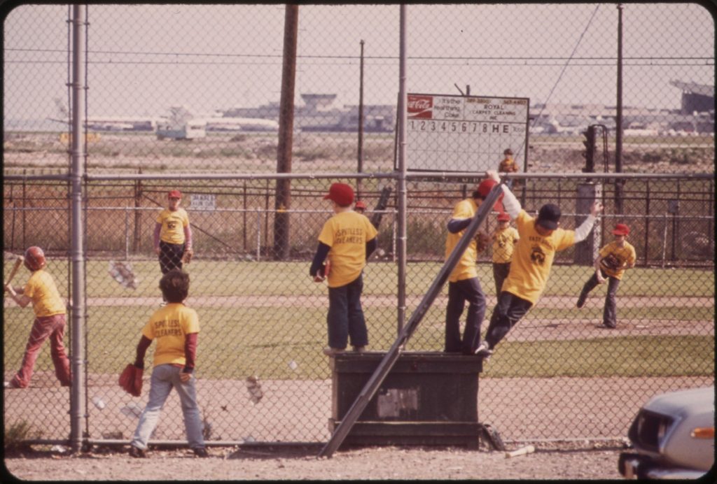 Boys in yellow jerseys and red caps play a Little League game behind a chain-link backstop, with airport buildings in the distance.