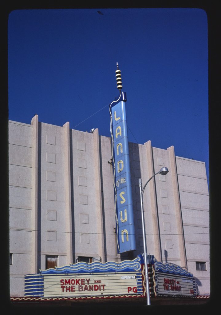 A tall pale theater with a blue vertical sign reading Land of the Sun and a marquee below reading Smokey and the Bandit, PG.