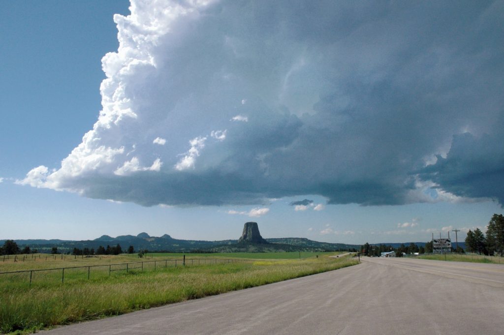 Devils Tower, a flat-topped rock column, rises on the horizon beyond a straight highway under a towering white storm cloud.