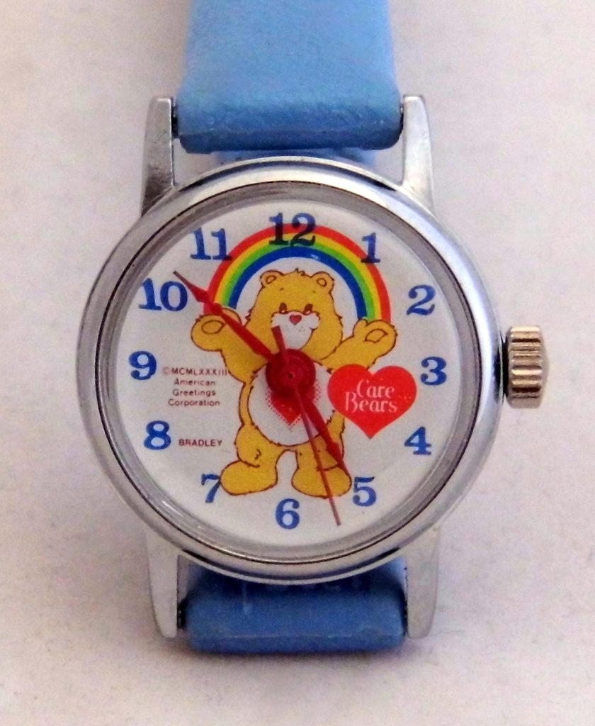 A child's wristwatch with a light blue strap and a yellow Care Bear under a rainbow on the dial, marked Bradley and American Greetings Corporation