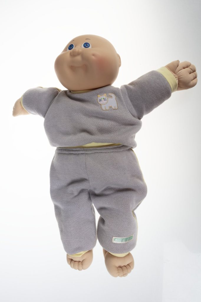 A bald Cabbage Patch Kids baby boy doll with blue eyes and a round vinyl face, wearing a gray track suit with yellow trim and a kitten patch