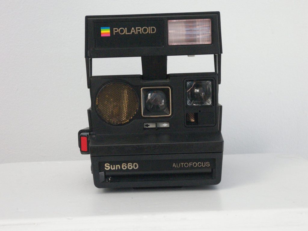 A black Polaroid Sun 660 Autofocus instant camera seen from the front, with a rainbow Polaroid logo, a flash bar on top and a gold round sensor beside the lens