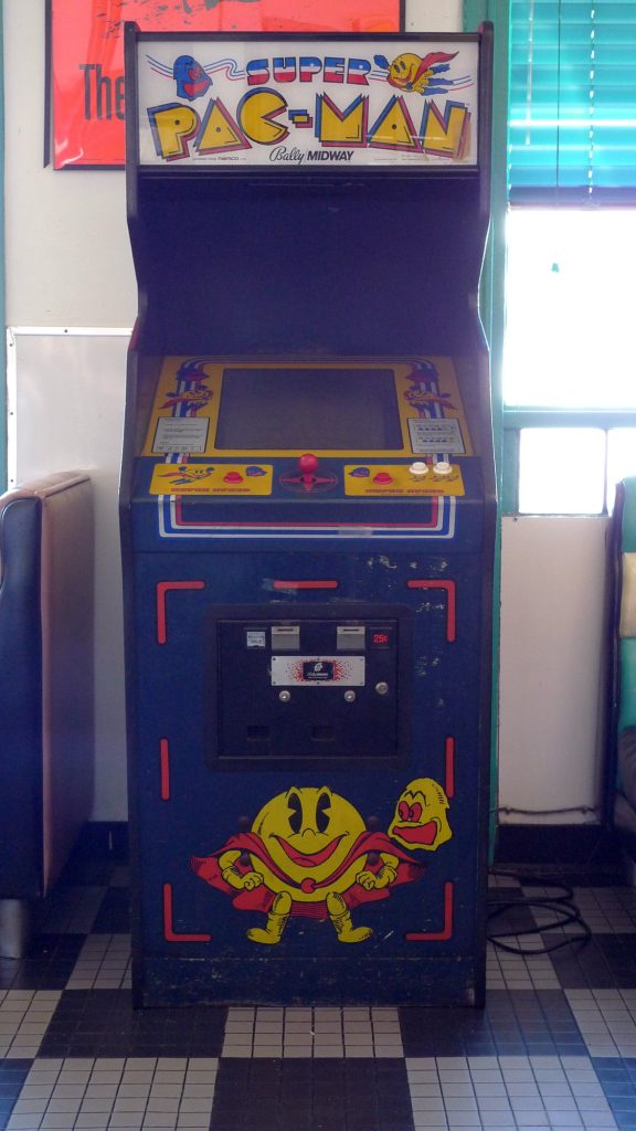A blue Super Pac-Man arcade cabinet by Bally Midway with a yellow control panel, a single red joystick and a caped Pac-Man painted on the front, standing on a checkered diner floor