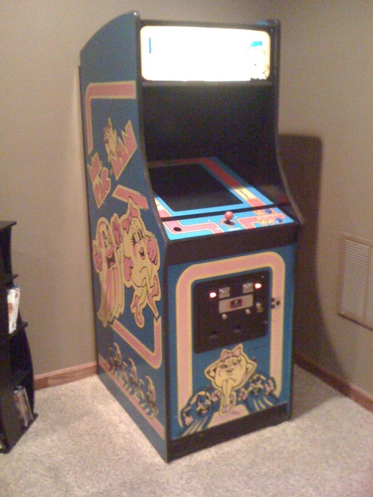 A blue Ms. Pac-Man upright arcade cabinet with pink and yellow cartoon artwork standing in the corner of a carpeted room