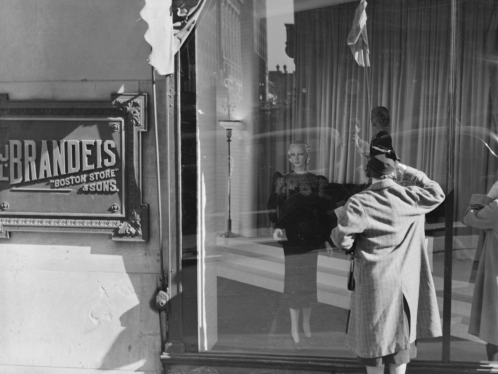 A woman in a plaid coat looks at dressed mannequins in the display window of the J.L. Brandeis & Sons “Boston Store,” beside the store’s name plaque.