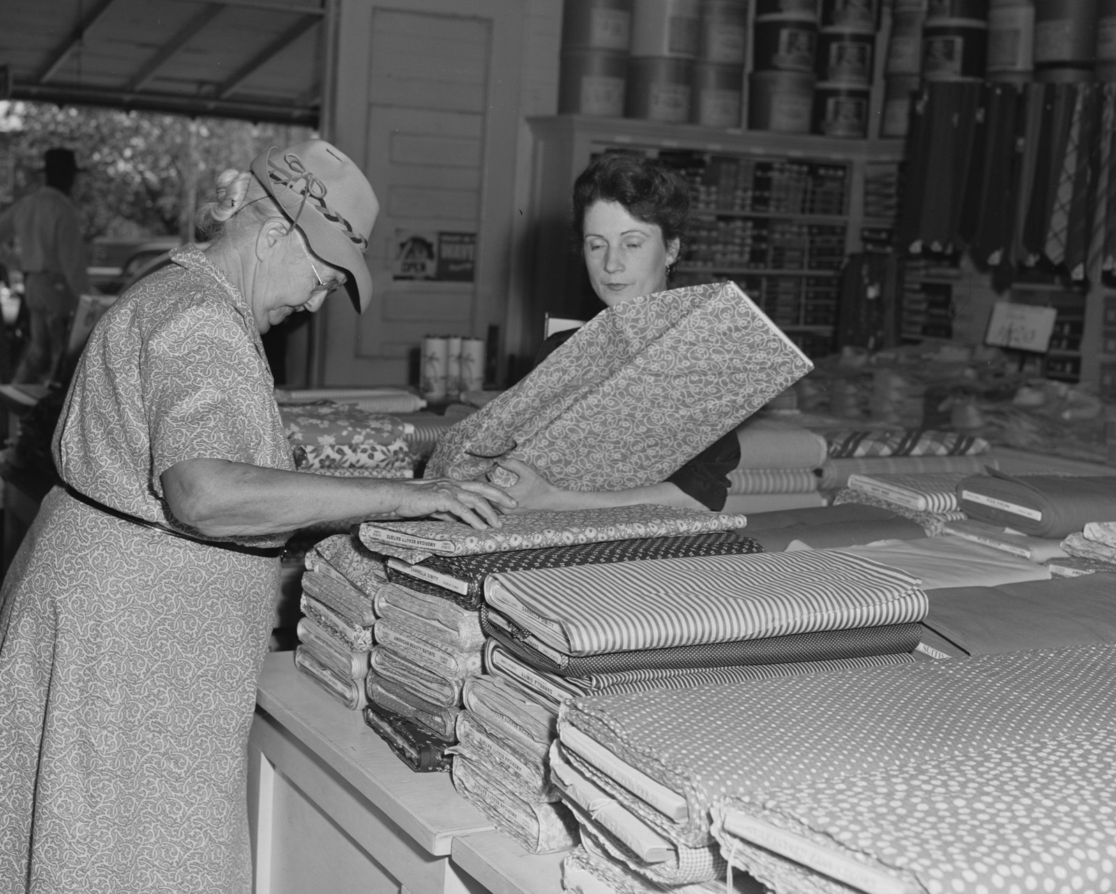 A woman in a hat examines folded bolts of patterned fabric on a store counter while a clerk holds up another bolt.