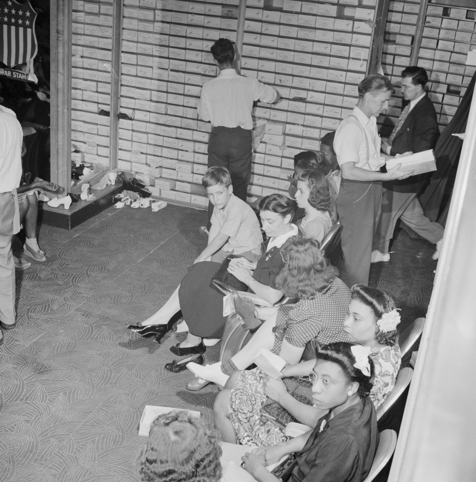 Customers sit in rows of chairs in a shoe store while salesmen pull boxes from floor-to-ceiling shelves.