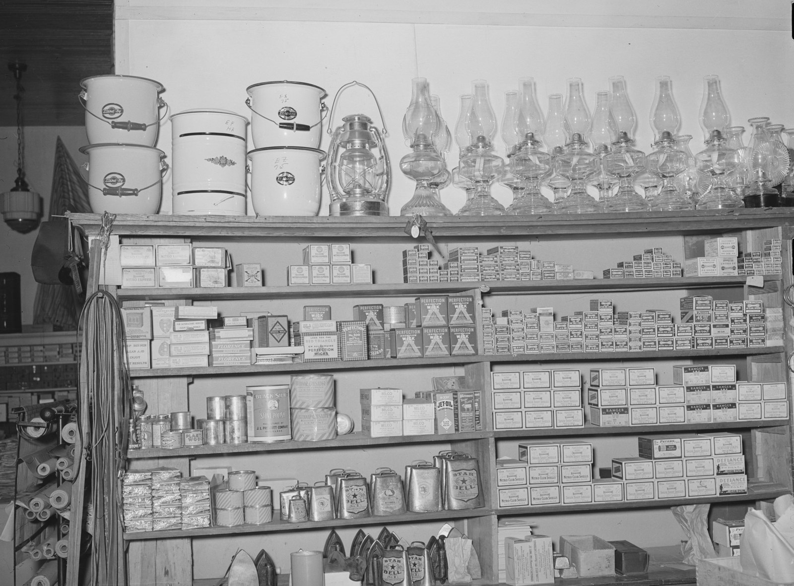 Shelves of boxed hardware with enamel pails and glass oil lamps lined up on the top shelf.