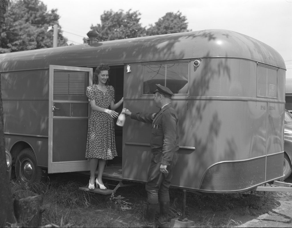 A uniformed milkman hands a glass quart bottle of milk to a smiling woman standing in the doorway of a house trailer