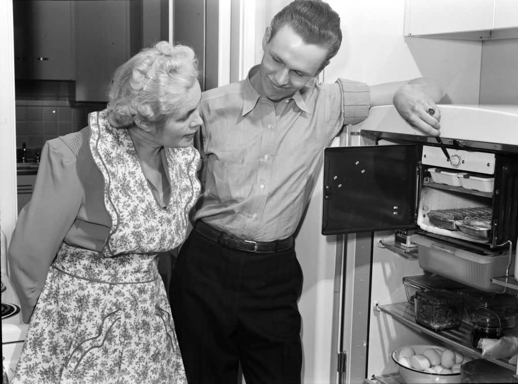 A woman and a young man look into a refrigerator whose small freezer box at the top holds frosted metal ice-cube trays