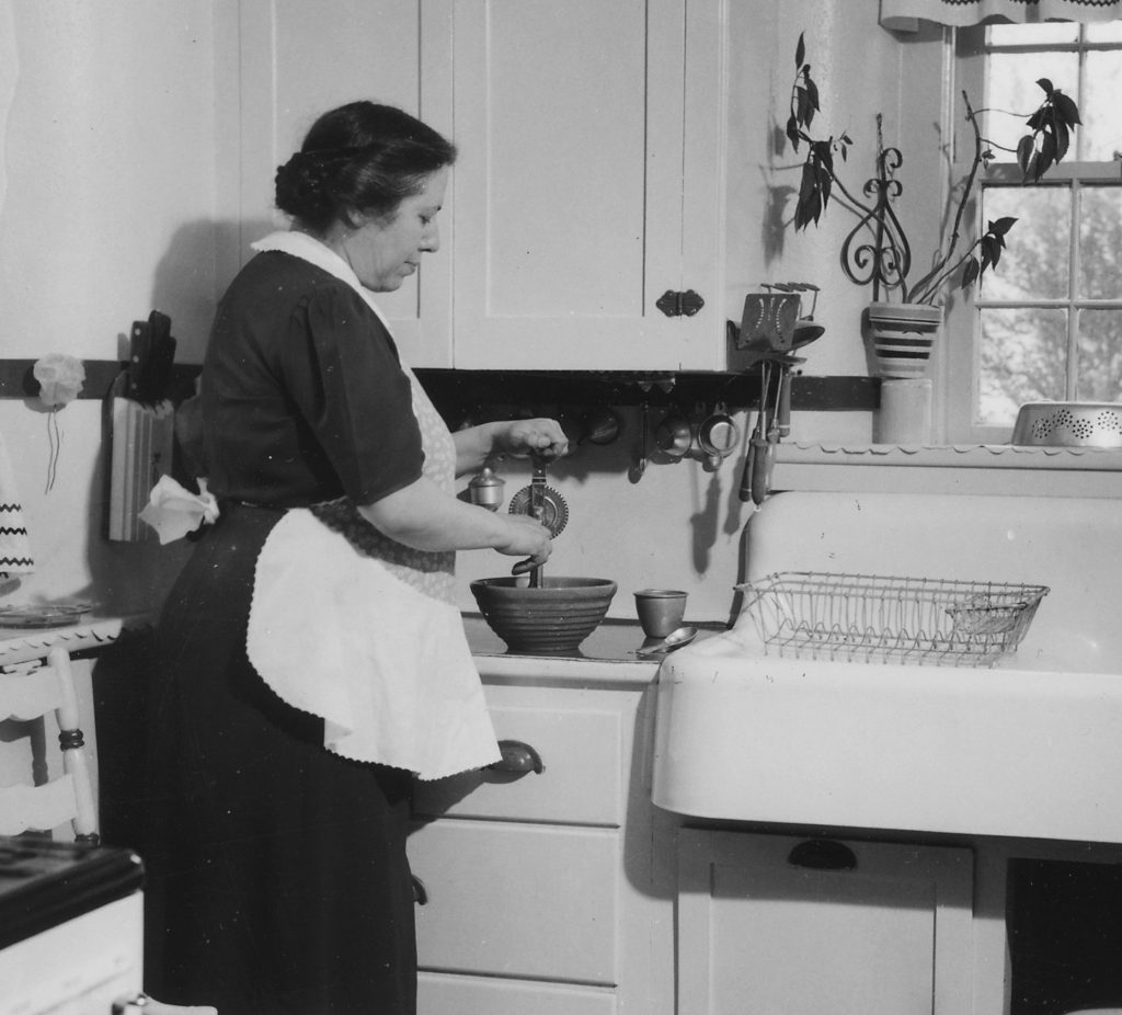 A woman in an apron turns the crank of a hand eggbeater in a stoneware mixing bowl beside a farmhouse sink