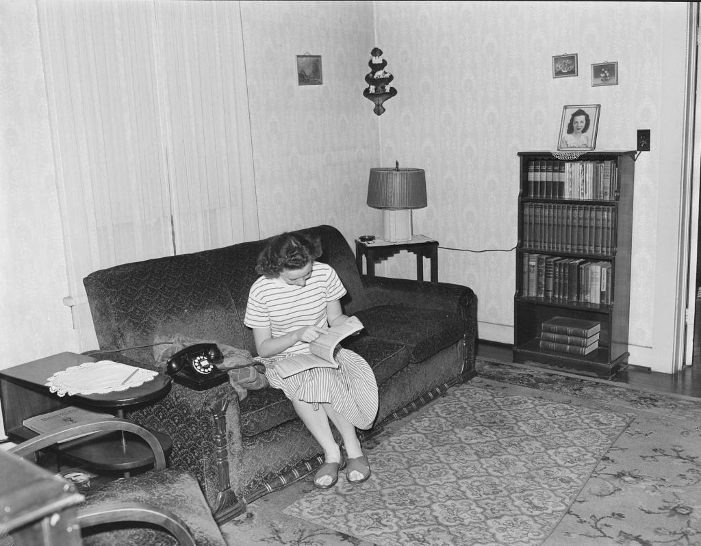 A young woman on a sofa looks up a number in a telephone directory, with a black rotary phone beside her