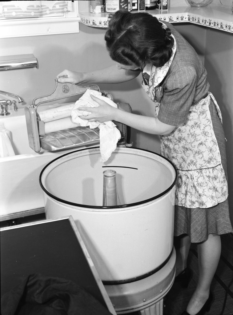 A woman wipes the rubber rolls of a wringer mounted on a round washing-machine tub