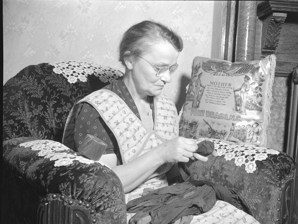 A gray-haired woman in glasses darns a dark sock with needle and thread, seated in an upholstered armchair