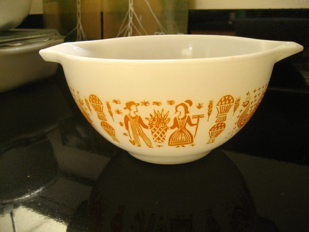 A white Pyrex Cinderella mixing bowl with pouring spouts, printed in gold with a farm couple, wheat sheaves and roosters