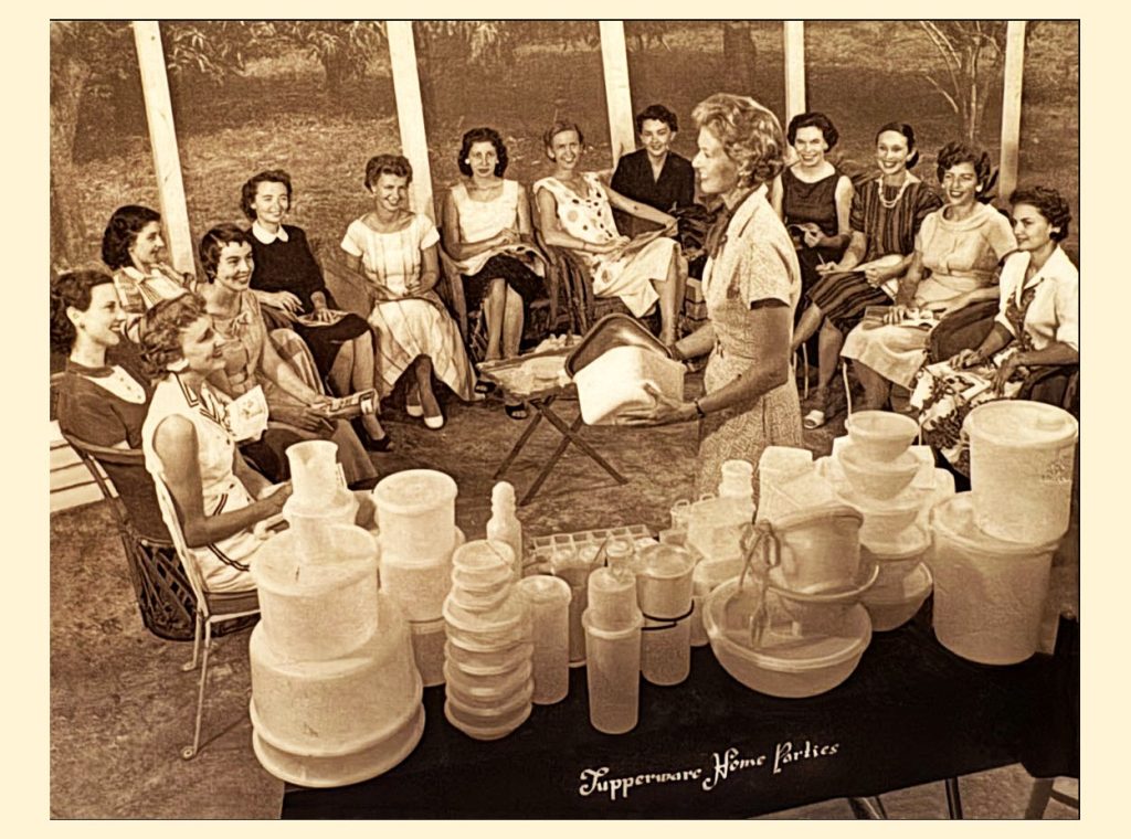 A hostess holds up a plastic container for a circle of seated women, with a table of Tupperware in front of her