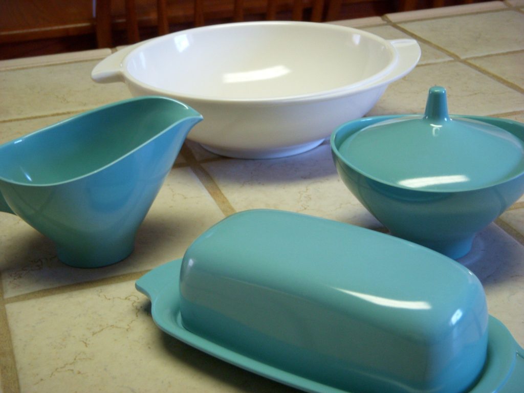 A turquoise melamine butter dish, creamer and lidded sugar bowl beside a white serving bowl on a tiled counter