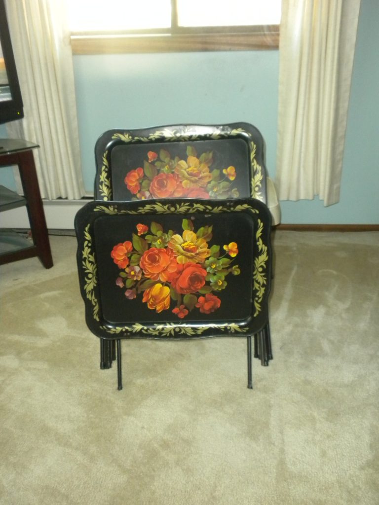 Black metal TV trays painted with orange and yellow roses, stacked in their folding stand on a carpet