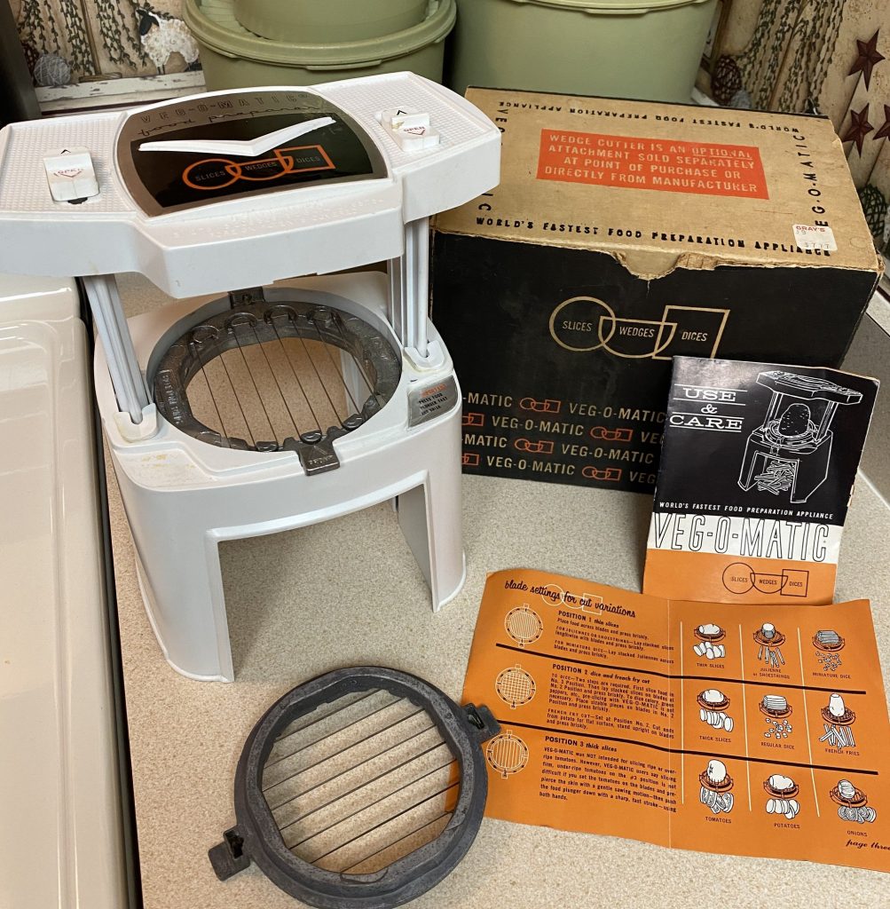 A white Veg-O-Matic slicer beside its black and orange box, with the instruction booklet and a spare cutting blade