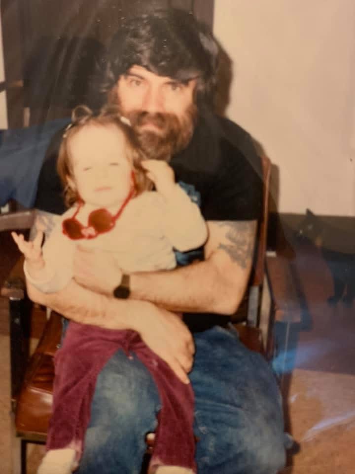 Jessica as a toddler, with red toy sunglasses around her neck, sitting on her bearded dad’s lap in the 1980s