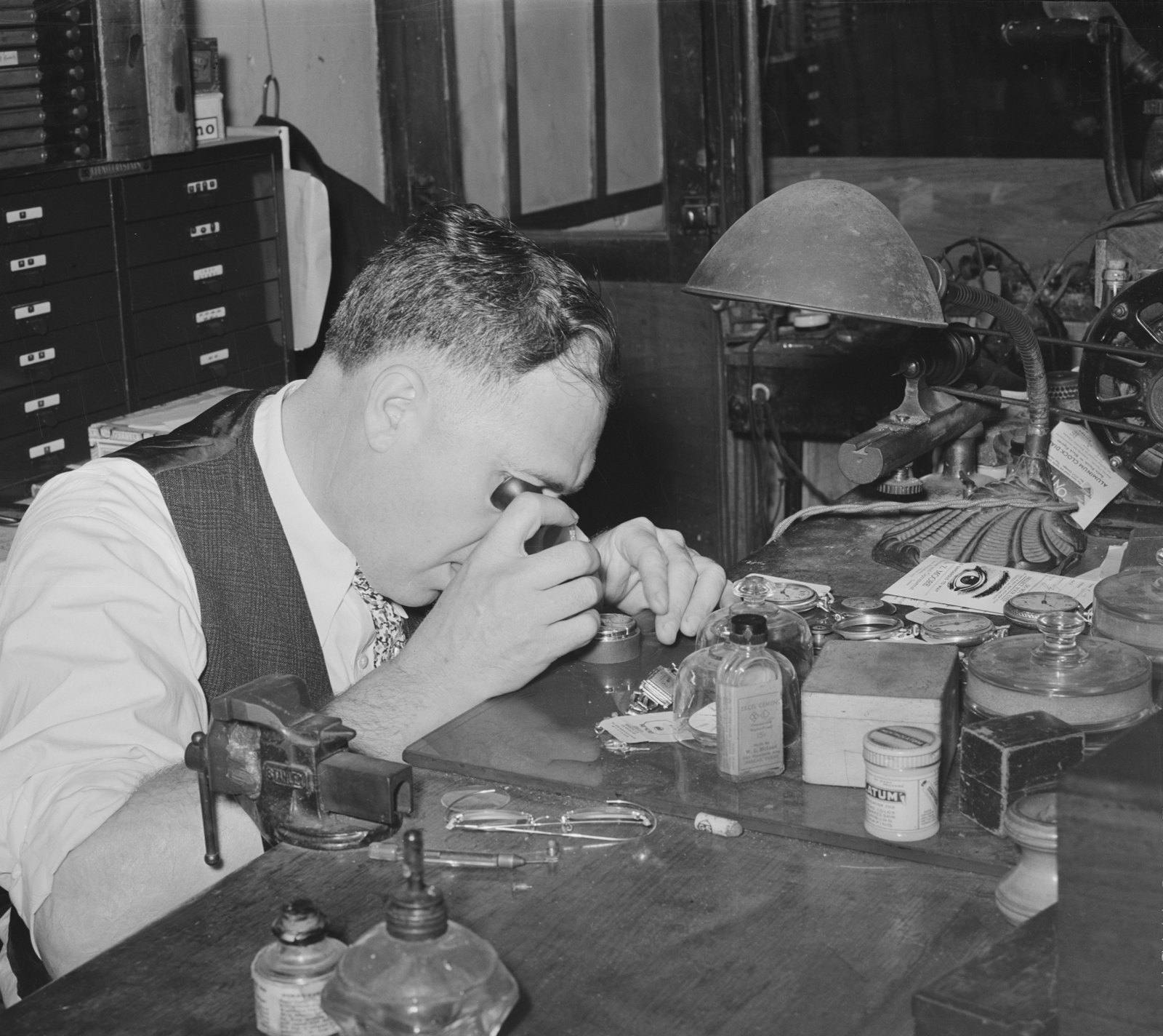 A jeweler wearing a loupe bends over his workbench to repair a watch.