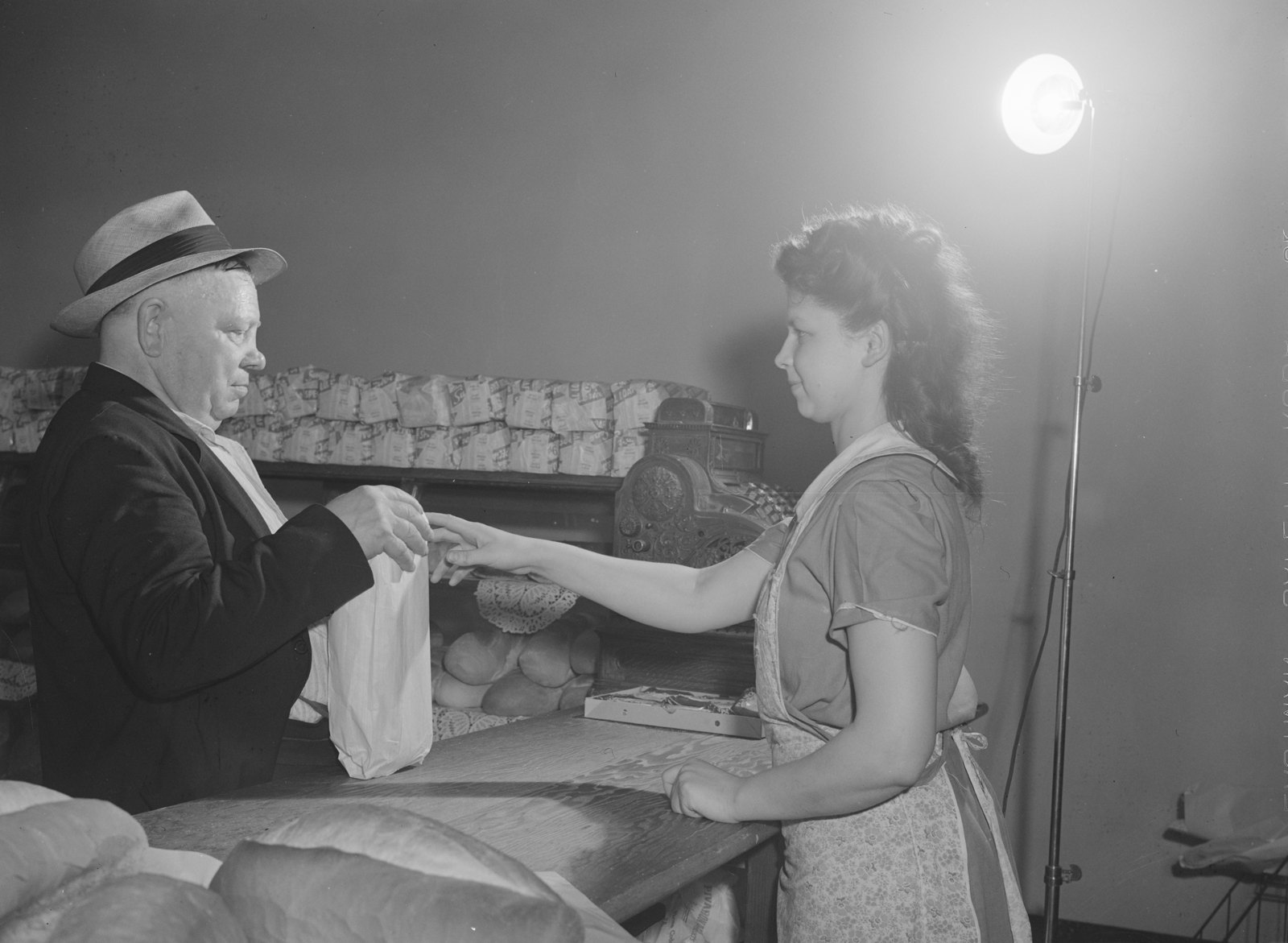 A customer in a straw hat hands money across a bakery counter to a clerk, with loaves of rye bread in the foreground.
