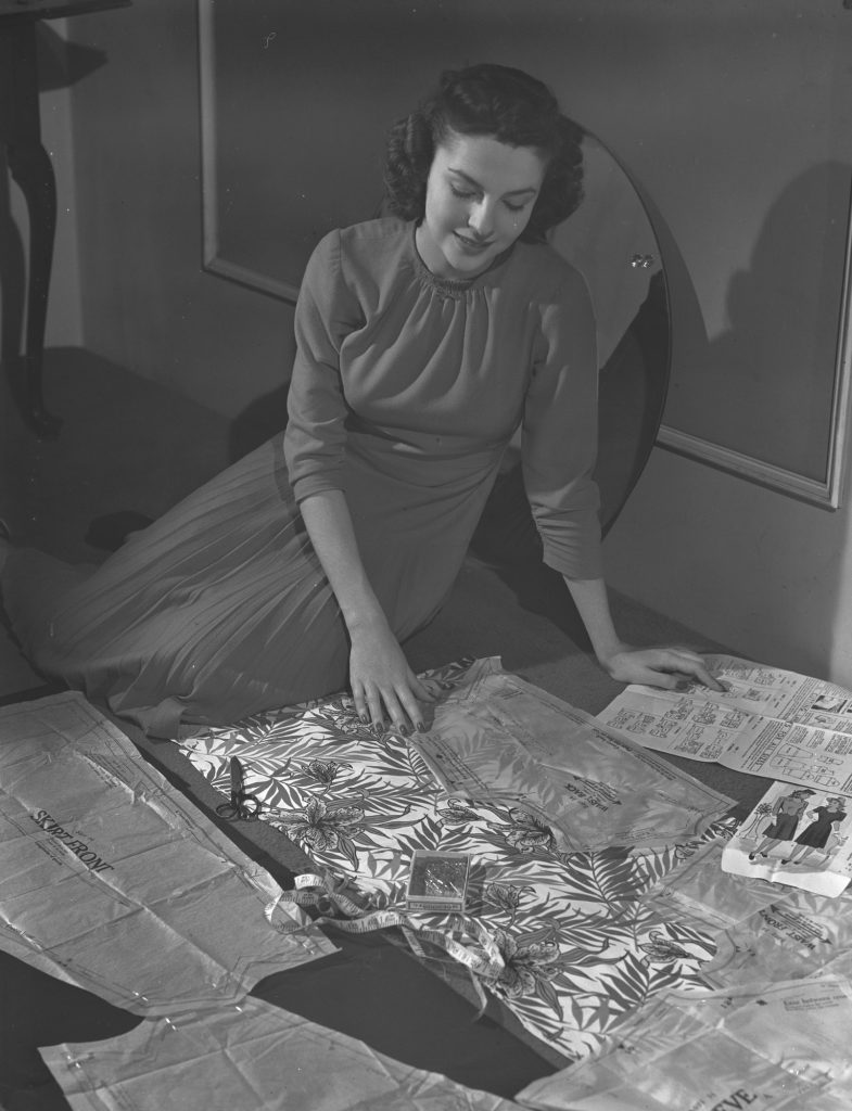 A smiling young woman kneels on the floor laying out tissue-paper dress pattern pieces on a length of leaf-print fabric, with scissors, a tape measure and the pattern envelope beside her