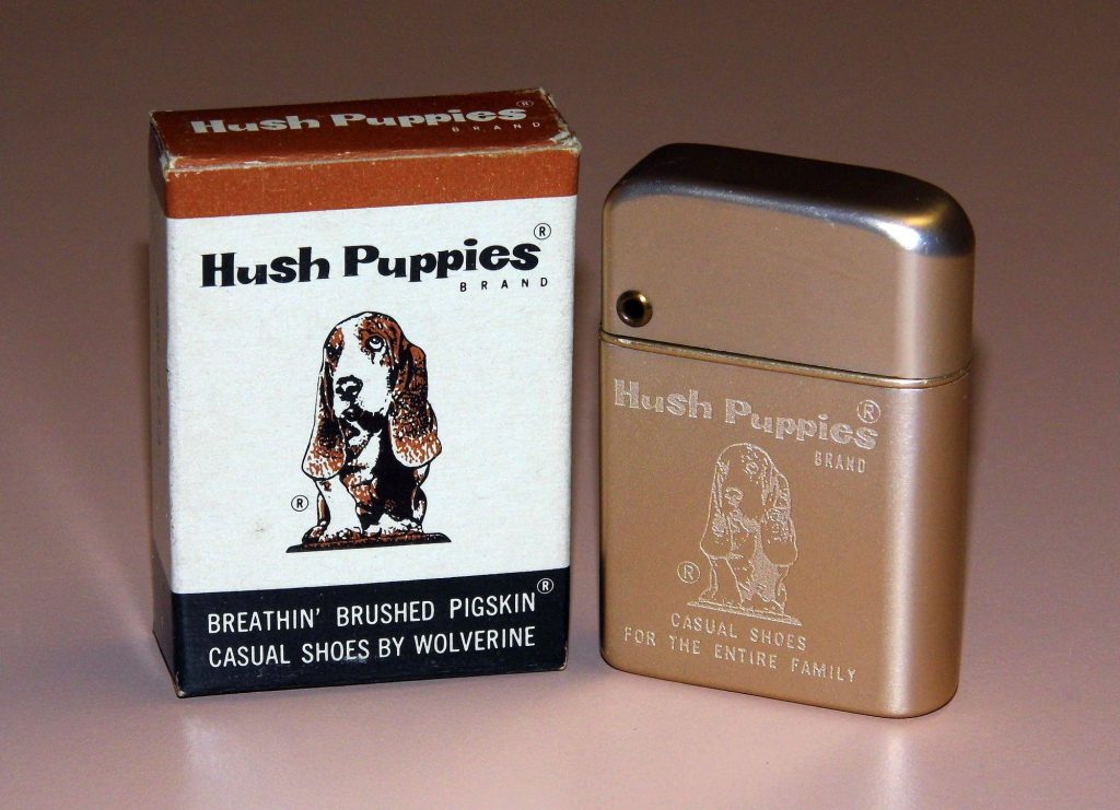 A Hush Puppies box printed with a sad-eyed basset hound and the words casual shoes by Wolverine, beside a gold cigarette lighter engraved with the same dog