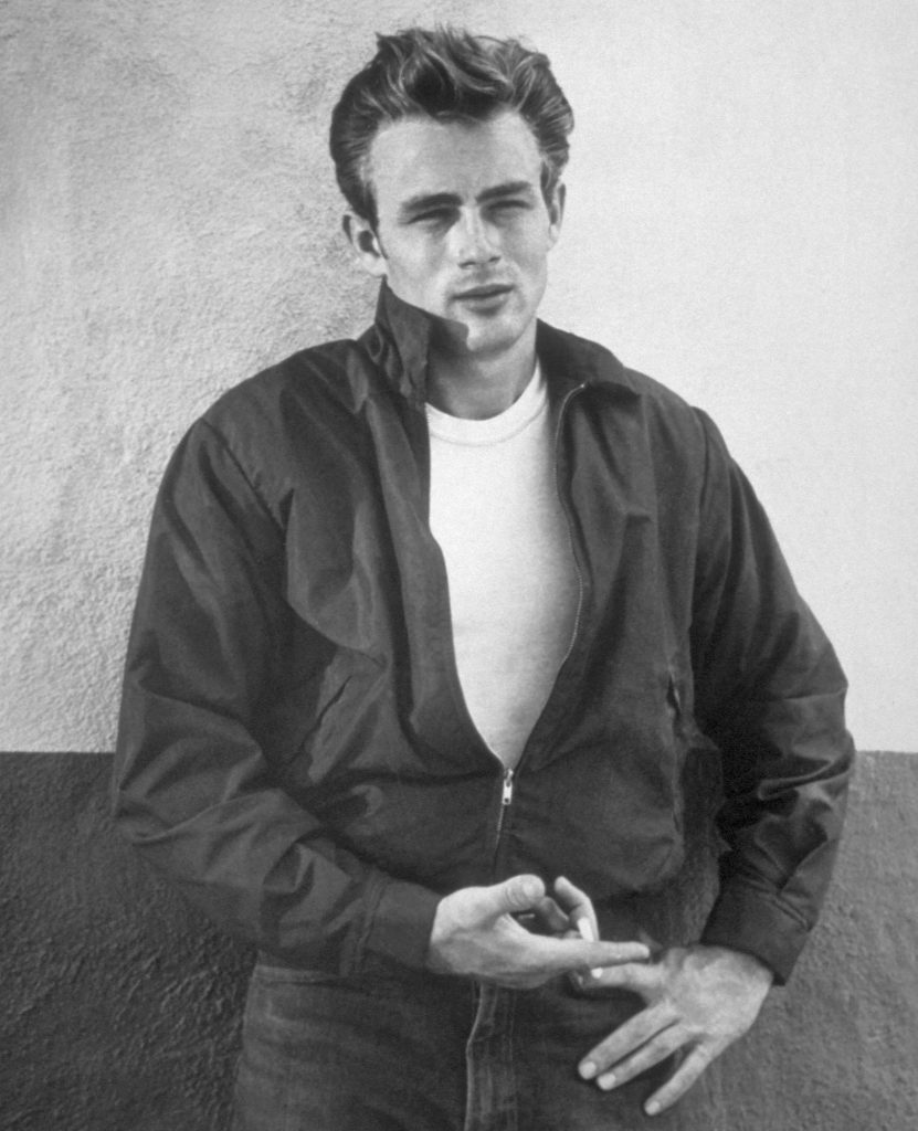 Black-and-white photo of James Dean leaning against a wall in a zip jacket, white T-shirt and jeans, holding a cigarette