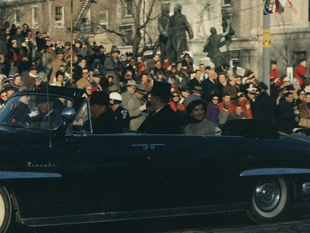 Jacqueline Kennedy smiling in a pale pillbox hat in the back of an open black Lincoln, beside President Kennedy in a black top hat, with crowds lining the street
