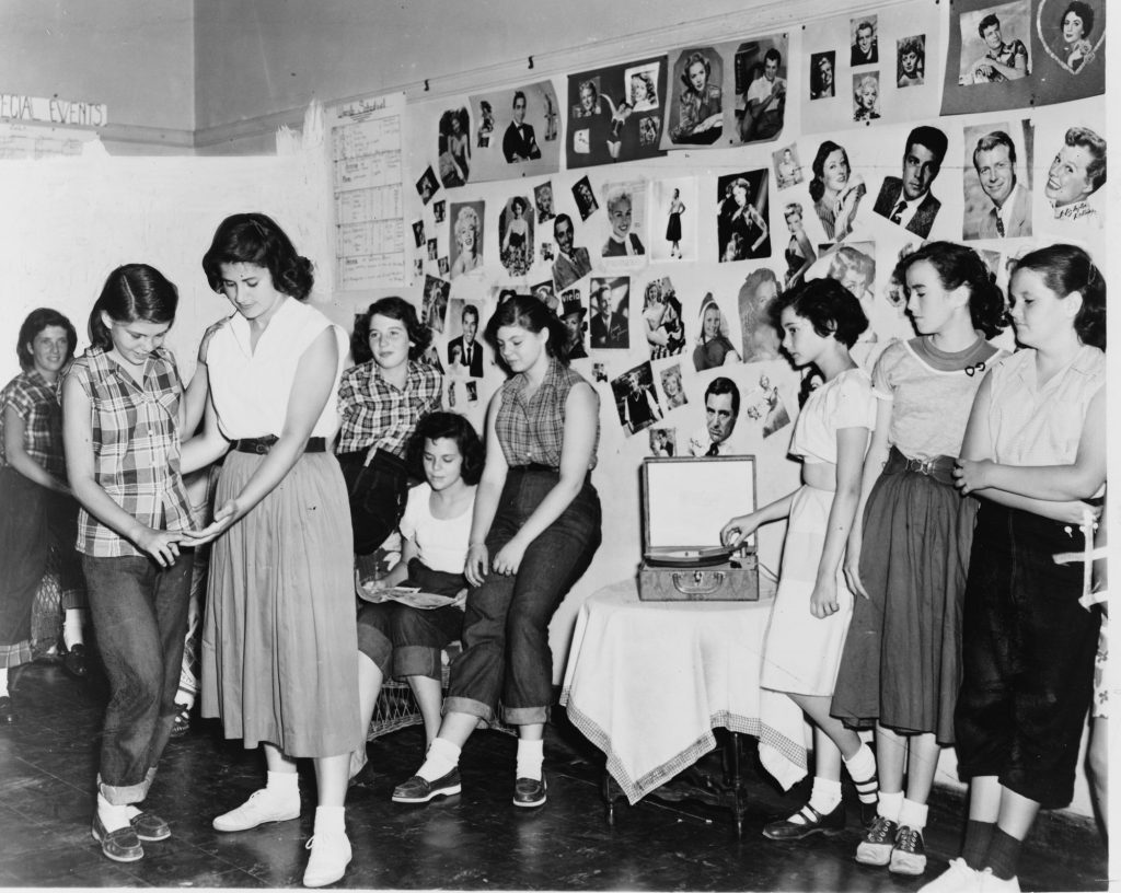 A row of teenage girls in full skirts, rolled jeans, bobby socks and saddle shoes, two of them practicing a dance step beside a portable record player, in front of a wall covered with movie-star photos