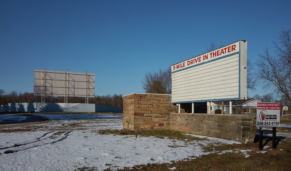 Snow-covered lot of the closed 5-Mile Drive In Theater, with a blank marquee on a stone base in front and the white screen in the distance