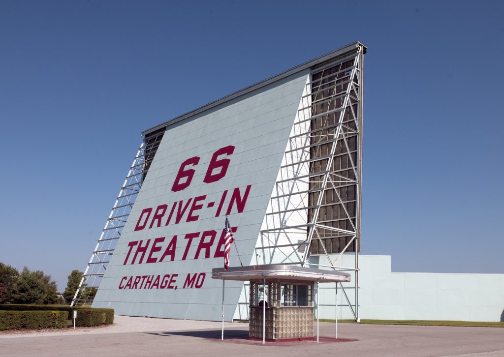 Tall white drive-in movie screen painted with 66 Drive-In Theatre Carthage, MO in red letters, with a small glass-block ticket booth and an American flag in front, under a clear blue sky