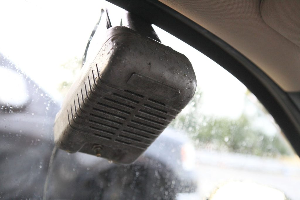 A dented gray metal drive-in speaker hooked over the rolled-down window of a car, seen from inside, with raindrops on the glass