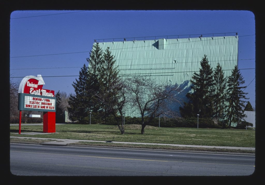 Red-and-white East Main drive-in sign with a marquee listing Redford and Fonda in Electric Horseman and Bruce Lee in Game of Death, in front of a tall pale green screen tower behind evergreen trees