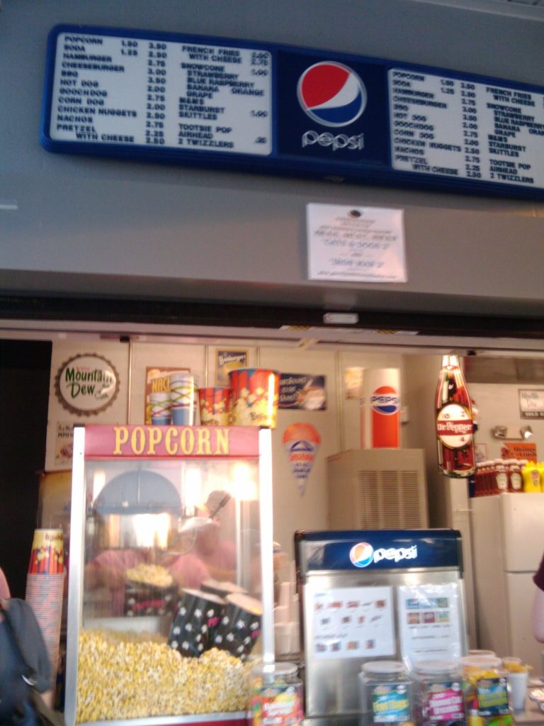 A drive-in snack bar counter with a glass popcorn machine full of popcorn, a Pepsi cooler, candy and soda signs, under a lighted menu board listing popcorn, hot dogs, nachos and drinks