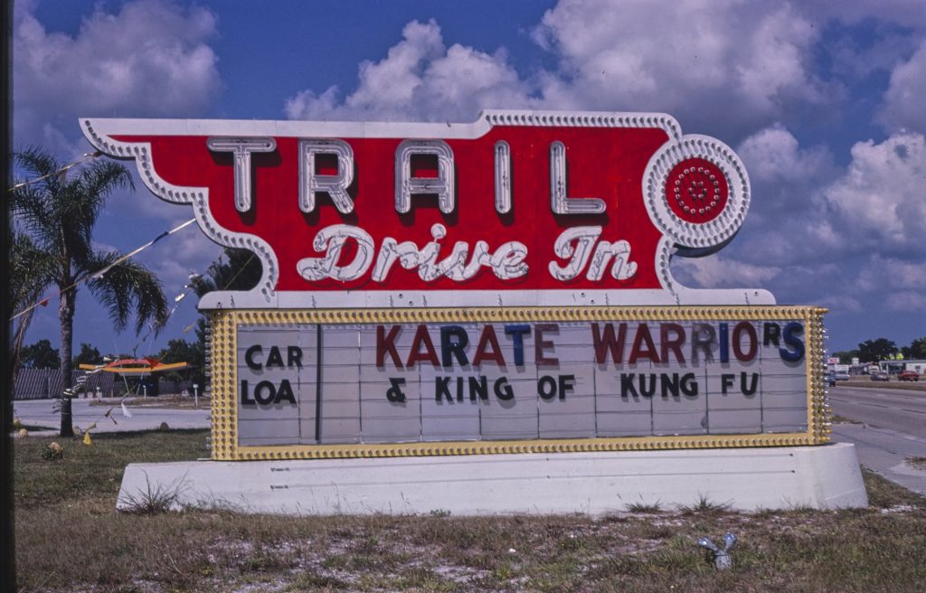 A red neon Trail Drive In sign over a marquee reading Car Loa, missing its final D, beside Karate Warriors and King of Kung Fu, with palm trees and a highway behind